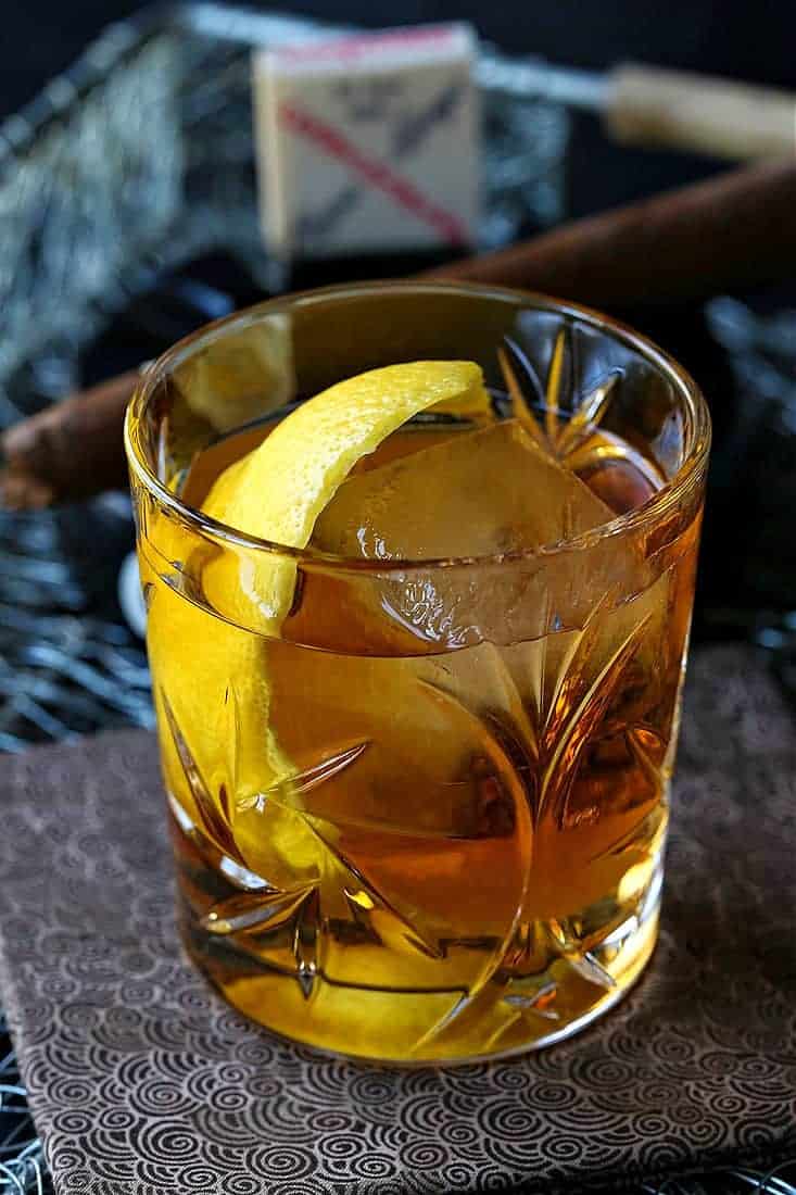 A bourbon cocktail in a rocks glass with ice and a lemon twist, with a cigar in the background