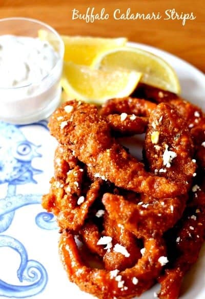 A pile of Buffalo Calamari Strips on a plate with lemons and blue cheese