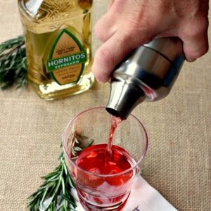 A bottle of reposado tequila on the counter next to a glass that someone is pouring cranberry drink from a cocktail shaker into.