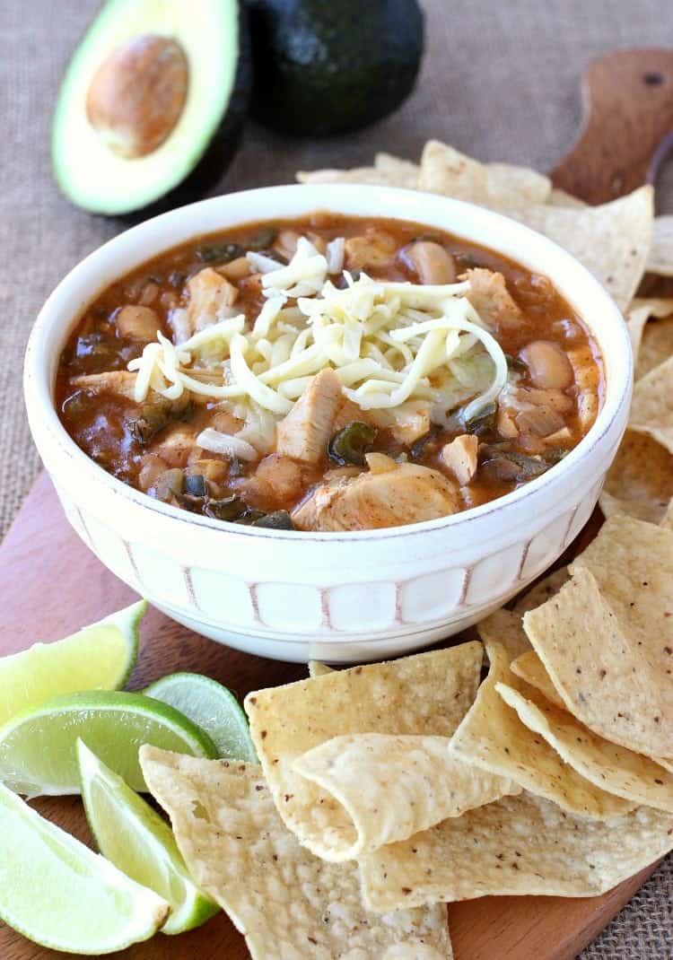 A bowl of white bean turkey chili topped with shredded cheese, surrounded by tortilla chips, lime wedges, and half an avocado