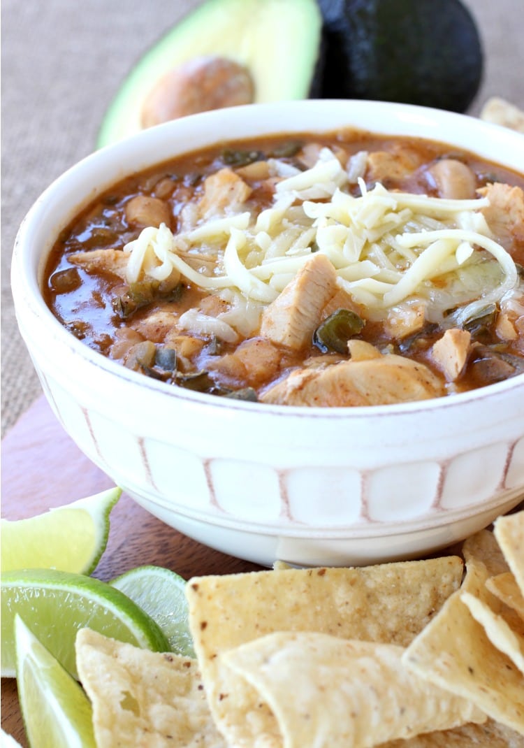 A bowl of white bean turkey chili topped with grated cheese, behind a pile of tortilla chips and lime wedges