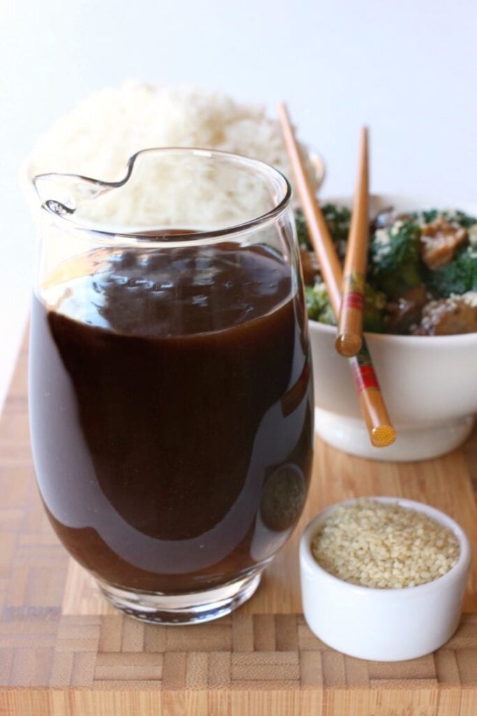 A big glass pitcher filled with homemade Stir Fry Sauce next to a bowl of sesame seeds