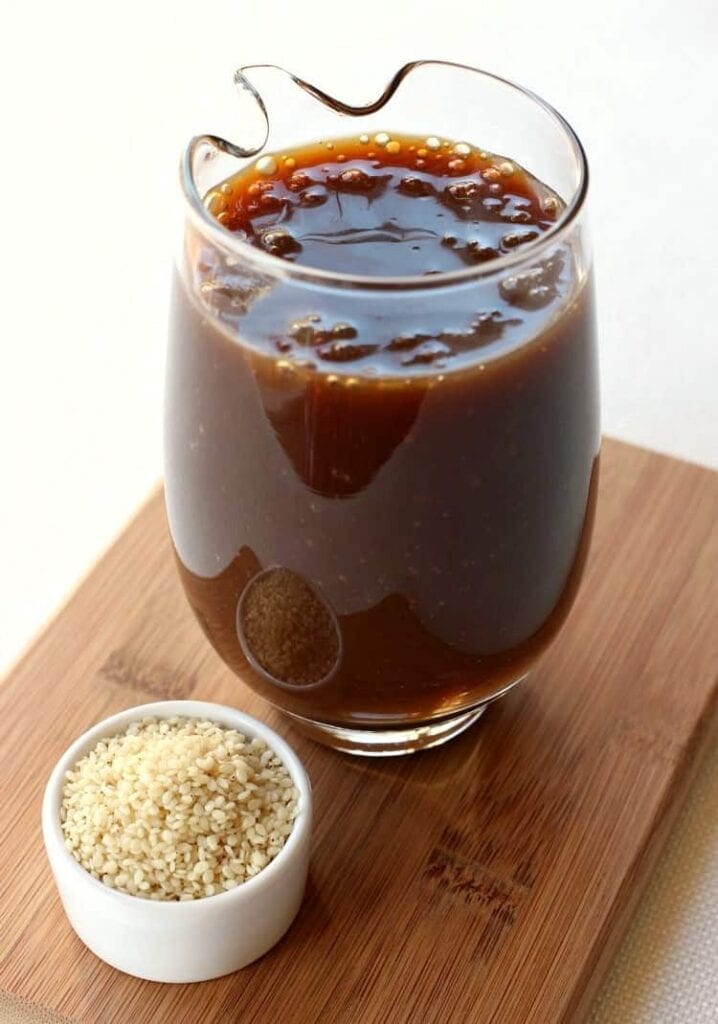 A glass cup filled with Stir Fry Sauce next to a bowl of sesame seeds on a wooden board