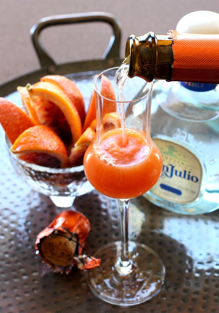 Champagne being poured into a cocktail with a bottle of tequila and a bowl of blood orange slices in the background