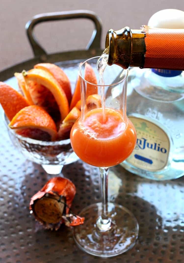 These 11 champagne cocktail recipes are perfect for a party, Christmas day, or a romantic night in, plus they're easy to make! Champagne being poured into a cocktail with a bottle of tequila and a bowl of blood orange slices in the background