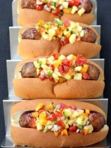 Four bratwurst sausages piled with Pineapple Tequila Salsa on a metal tray.