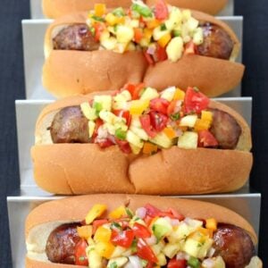 Four bratwurst sausages piled with Pineapple Tequila Salsa on a metal tray.