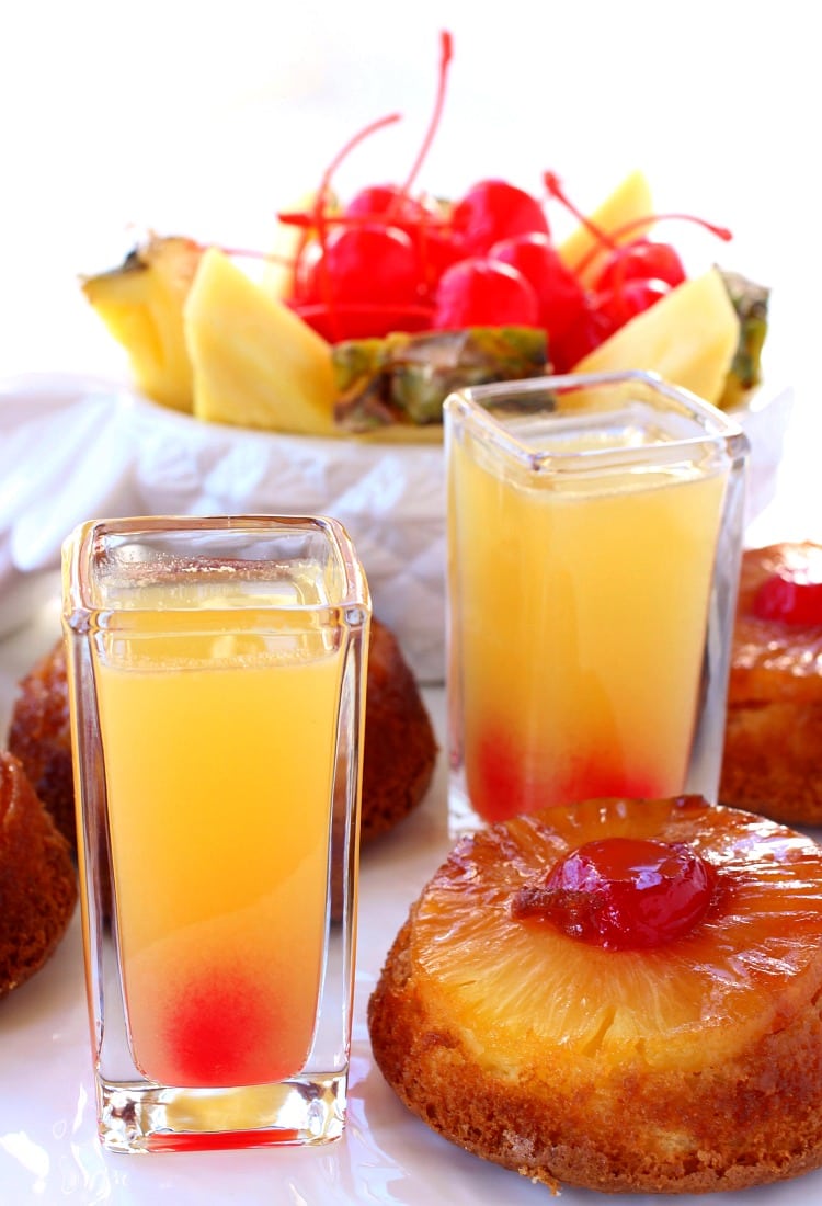 Two pineapple upside down shots surrounded by pieces of pineapple upside down cake, with a bowl of pineapples and maraschino cherries in the background