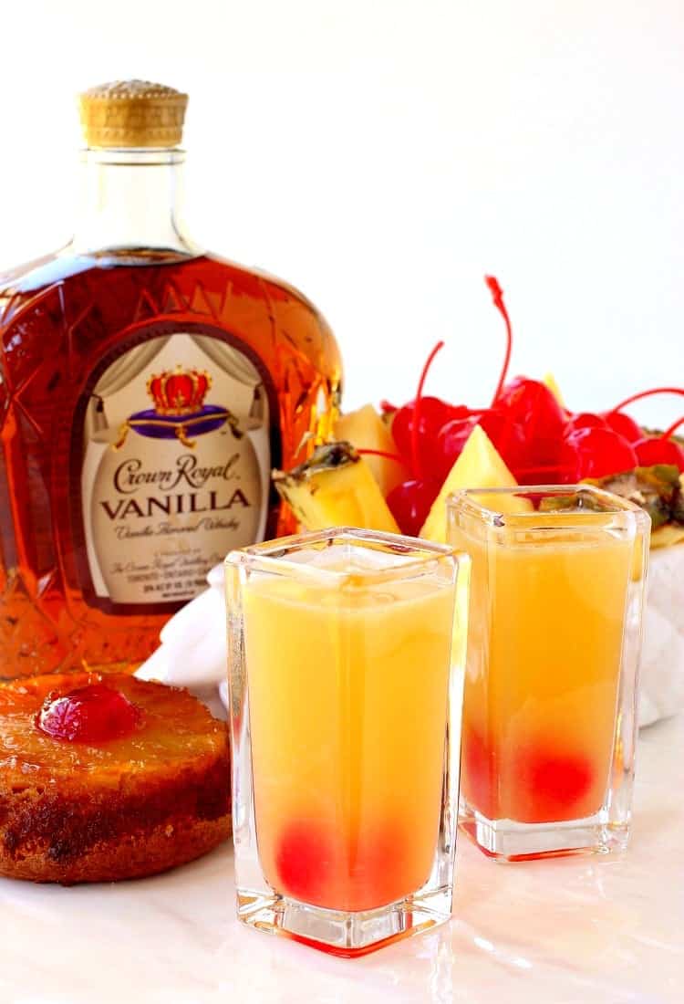 Two pineapple upside down shots next to a slice of pineapple upside down cake, a bottle of Crown Royal vanilla whiskey, and a down of pineapple slices and maraschino cherries