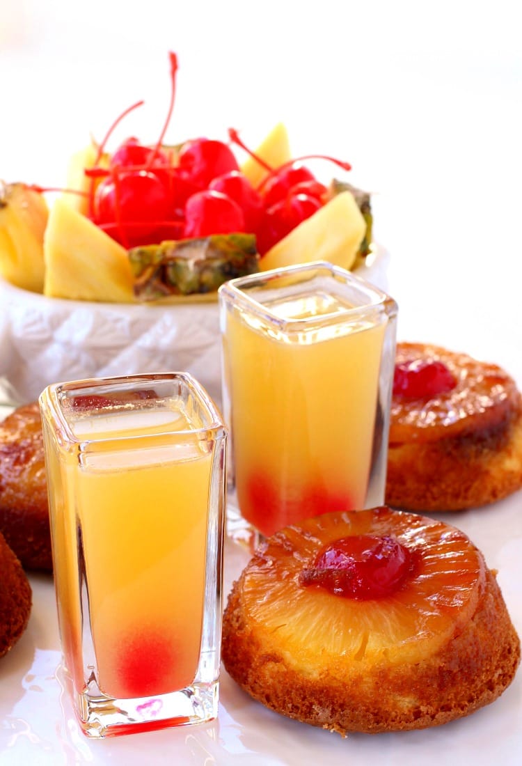 Two pineapple upside down shots next to a few slices of pineapple upside down cake, with a bowl of pineapple slices and maraschino cherries in the background 