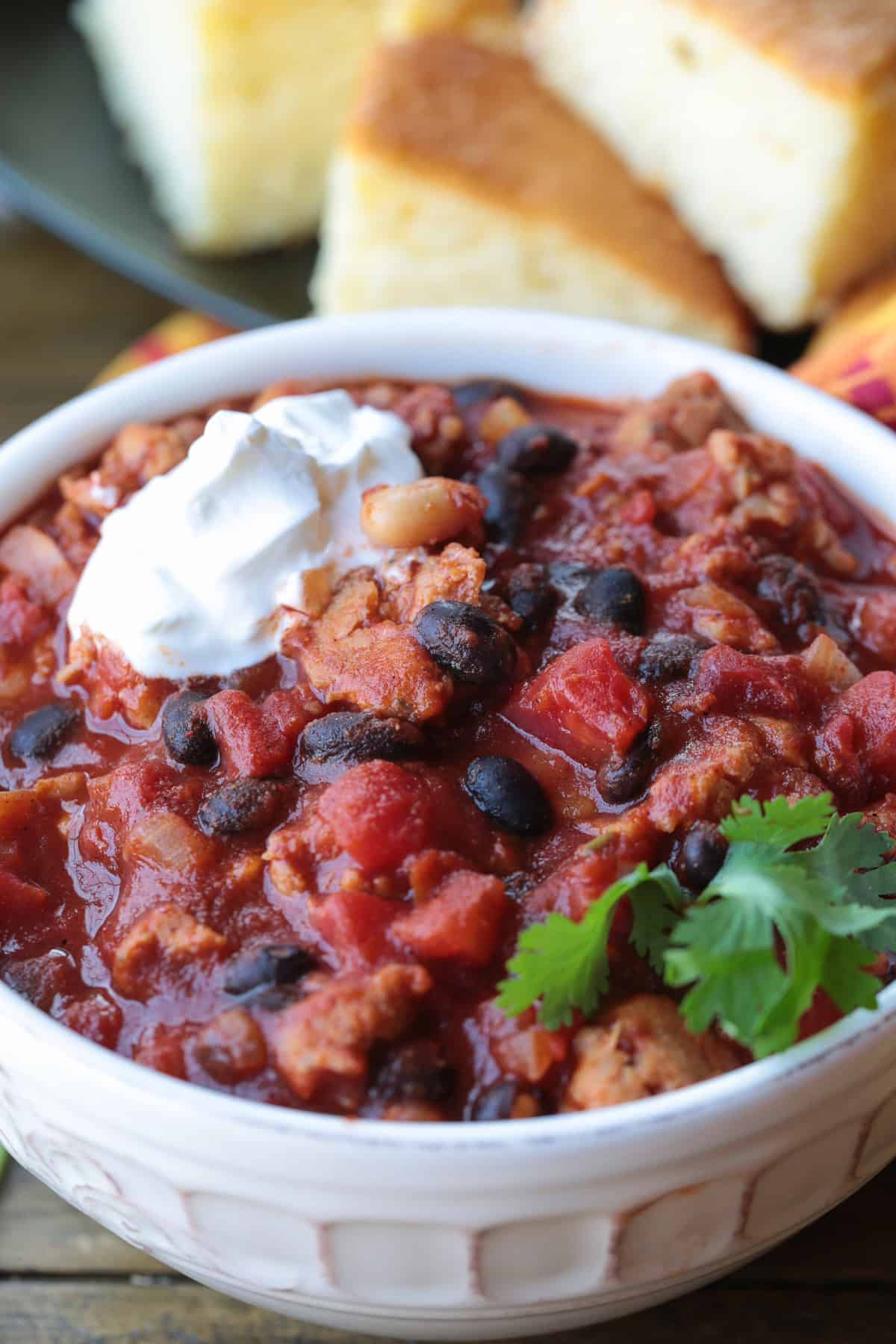 This Chicken Sausage Chili is super hearty and packed with flavor! We like a combo of sweet and spicy sausage, lots of beans and bold spices. chili in a bowl with cornbread in the background