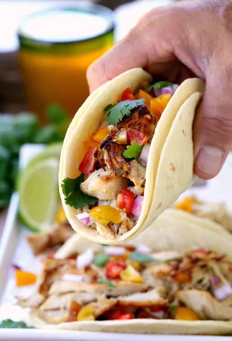 A hand holding a chicken taco with two tortillas, fresh veggies, and cilantro, with more tacos in the background