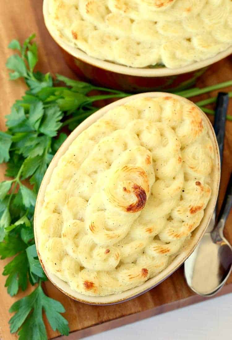 Overhead view of a turkey shepherd's pie with lightly browned mashed potatoes, next to another turkey shepherd's pie, some sprigs of parsley, and two spoons