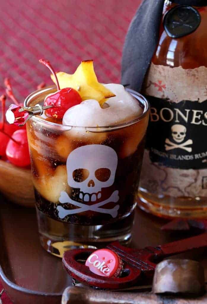 Rum and Coke in a glass with ice next to a bottle of rum and a bowl of maraschino cherries.