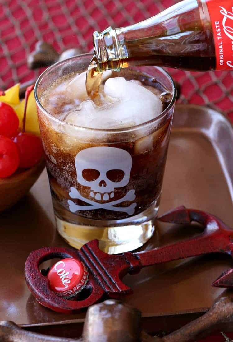 With a kick of cinnamon and dazzling flakes of real gold, this Rum and Coke cocktail gives the classic drink a decadent tropical twist. Coke being poured from a glass bottle into a glass with rum and Goldschlager.