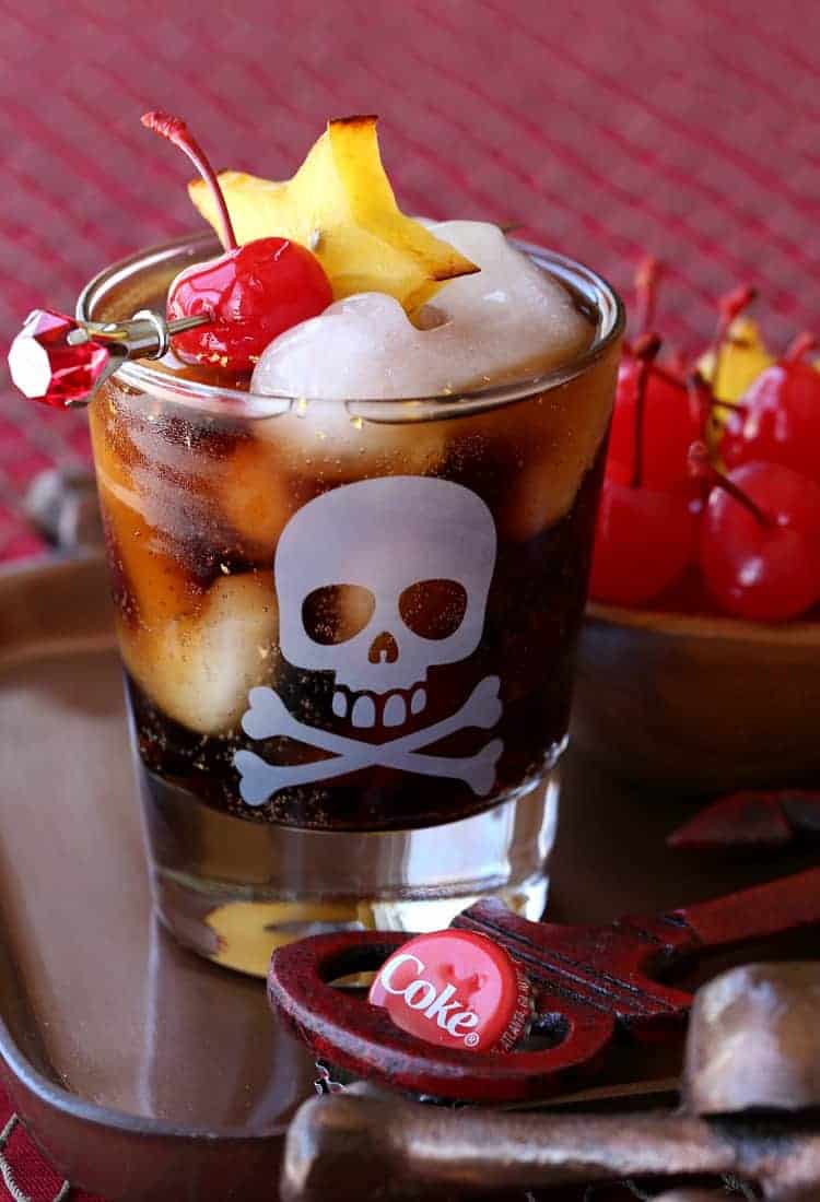 With a kick of cinnamon and dazzling flakes of real gold, this Rum and Coke cocktail gives the classic drink a decadent tropical twist. A glass of Rum and Coke garnished with a maraschino cherry and a slice of star fruit.