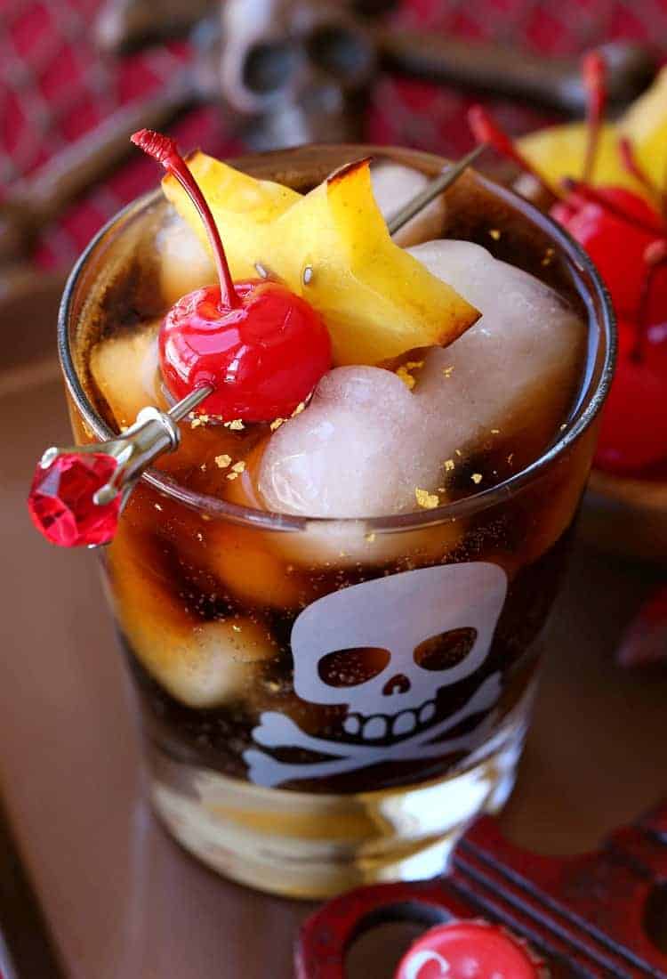 With a kick of cinnamon and dazzling flakes of real gold, this Rum and Coke cocktail gives the classic drink a decadent tropical twist. The top view of a glass filled with ice and bubbly cocktail with fresh fruit for garnish.