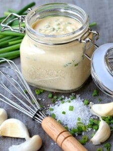 A jar of Awesome Sauce and a whisk next to scattered chives, garlic cloves, and salt