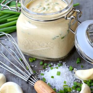 A jar of Awesome Sauce and a whisk next to scattered chives, garlic cloves, and salt