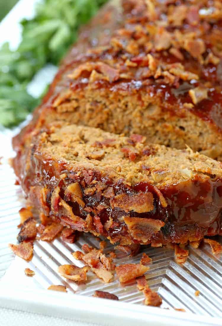 A slice of turkey meatloaf on a baking sheet, with the rest of the meatloaf behind it, and crumbled bacon around it