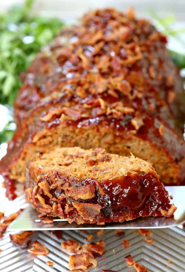 A slice of turkey meatloaf on a spatula, with the rest of the meatloaf in the background, surrounded by bacon bits and parsley