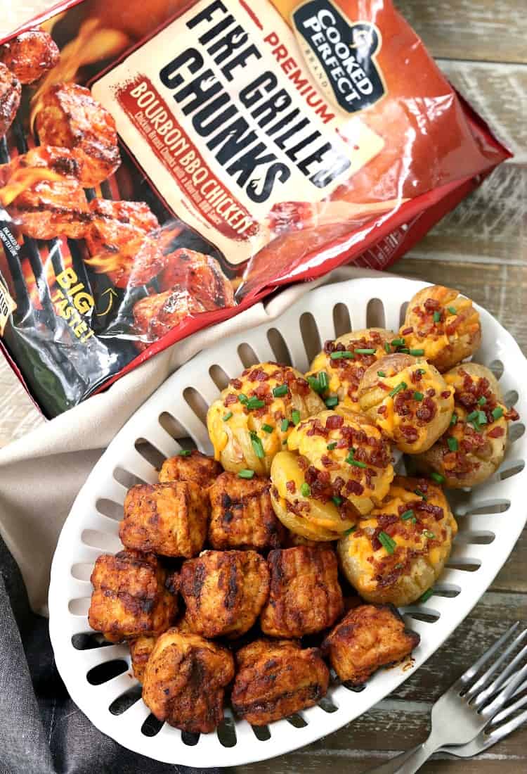A tray that is half full of mini loaded hasselback potatoes, and half full of chicken bites, surrounded by forks and a bag of chicken bites