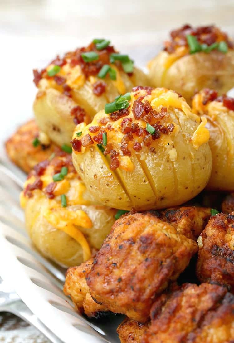 A tray of loaded hasselback potatoes topped with bacon bits, cheese, and chives, next to bite-size pieces of chicken