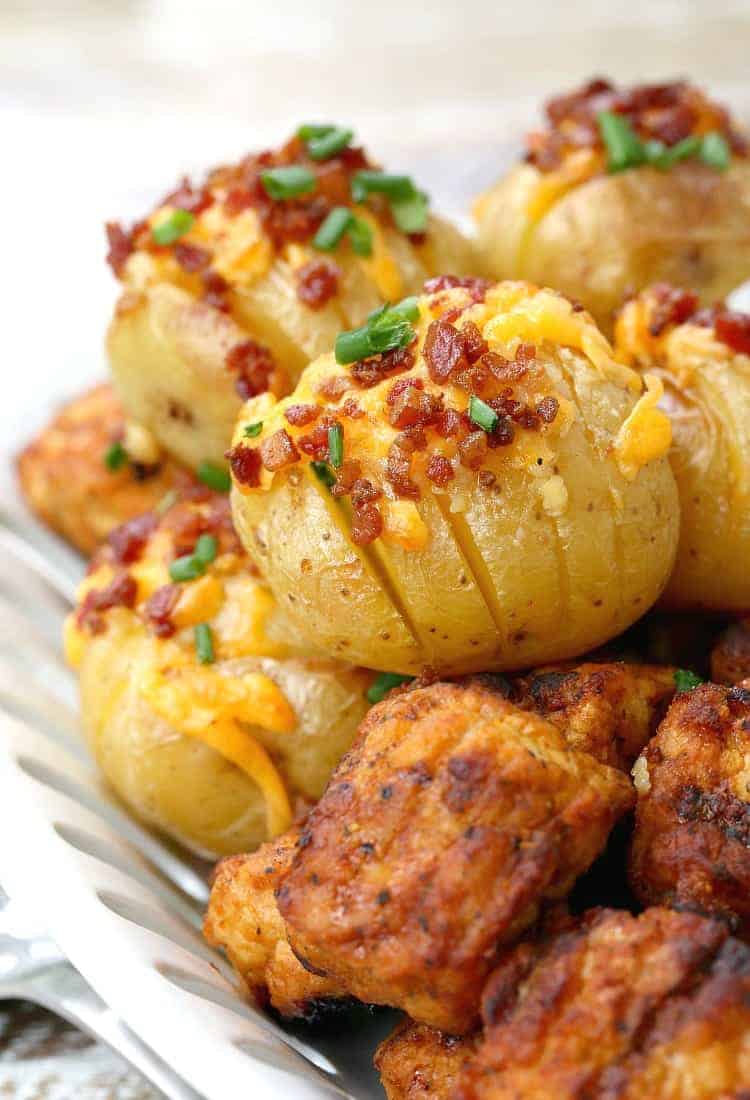 These mini loaded hasselback potatoes are full of gooey cheese and rich bacon, plus they're a quick and easy side dish to make A tray of loaded hasselback potatoes topped with bacon bits, cheese, and chives, next to bite-size pieces of chicken