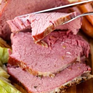 Sliced Crock Pot Corned Beef on a wooden cutting board next to braised cabbage and carrots.