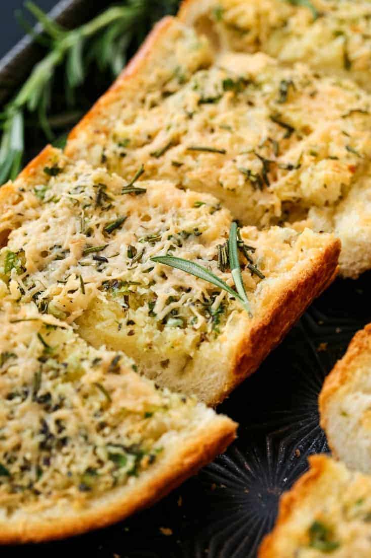 Rosemary Asiago Garlic Bread is herby and buttery with a crisp crust. Serve this garlic bread with everything from pasta to steak! Rosemary Asiago Garlic Bread sliced on a baking sheet with rosemary garnish