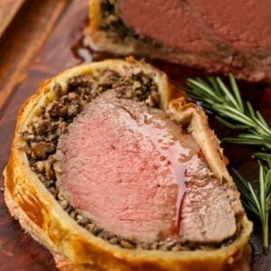 A slice of juicy beef wellington on a cutting board with sprigs of fresh rosemary.