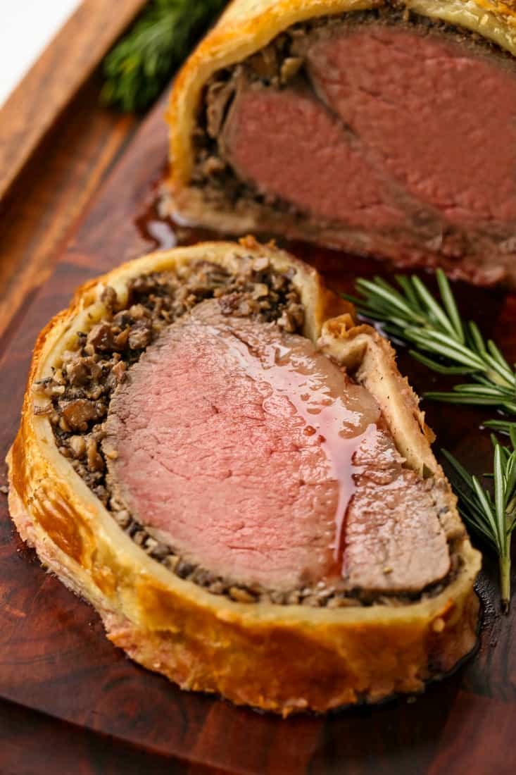 A slice of juicy beef wellington on a cutting board with sprigs of fresh rosemary.