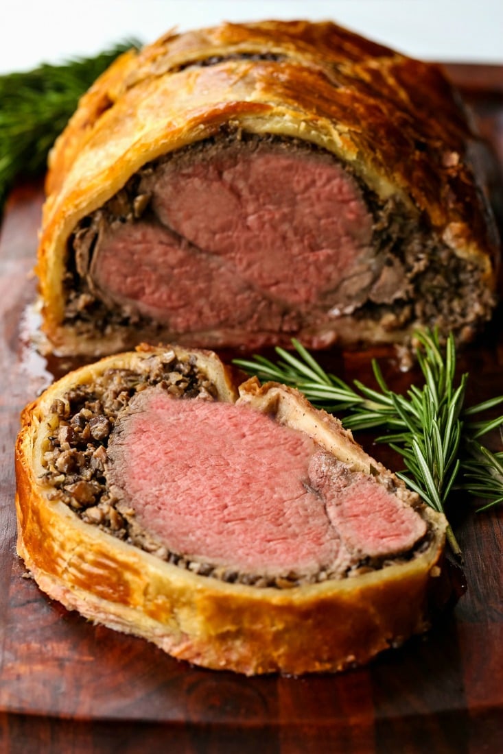 A beef wellington that's been cut into, with a slide on the cutting board, and rosemary surrounding it