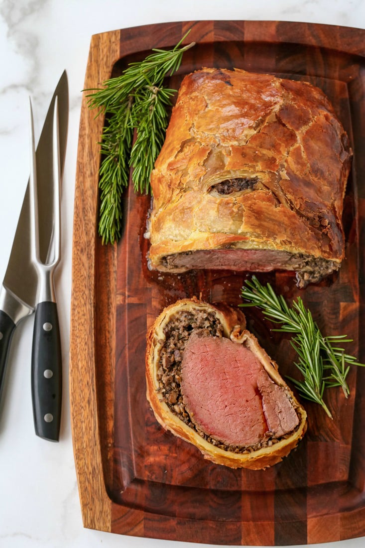 A slice of beef wellington on a board beside the remaining pastry-wrapped tenderloin.