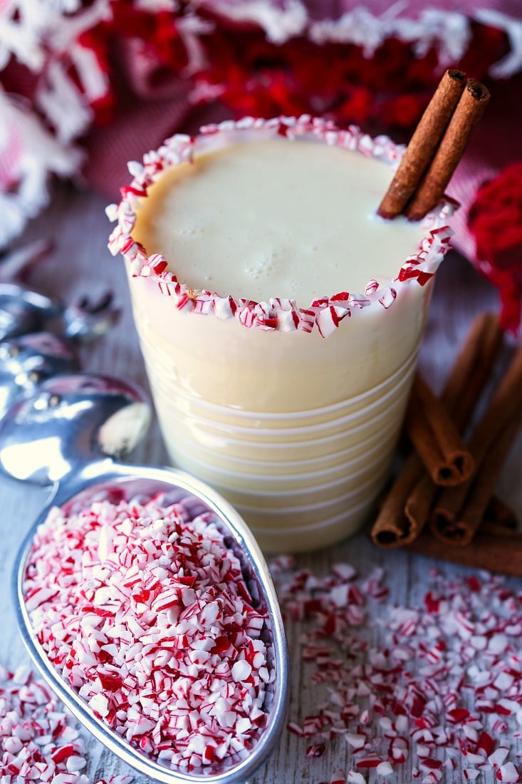 A peppermint eggnog cocktail in a glass, with crushed candy canes on the rim of the glass and a cinnamon stick for a straw, surrounded by candy cane chunks and cinnamon sticks, and a scoop full of candy cane pieces 