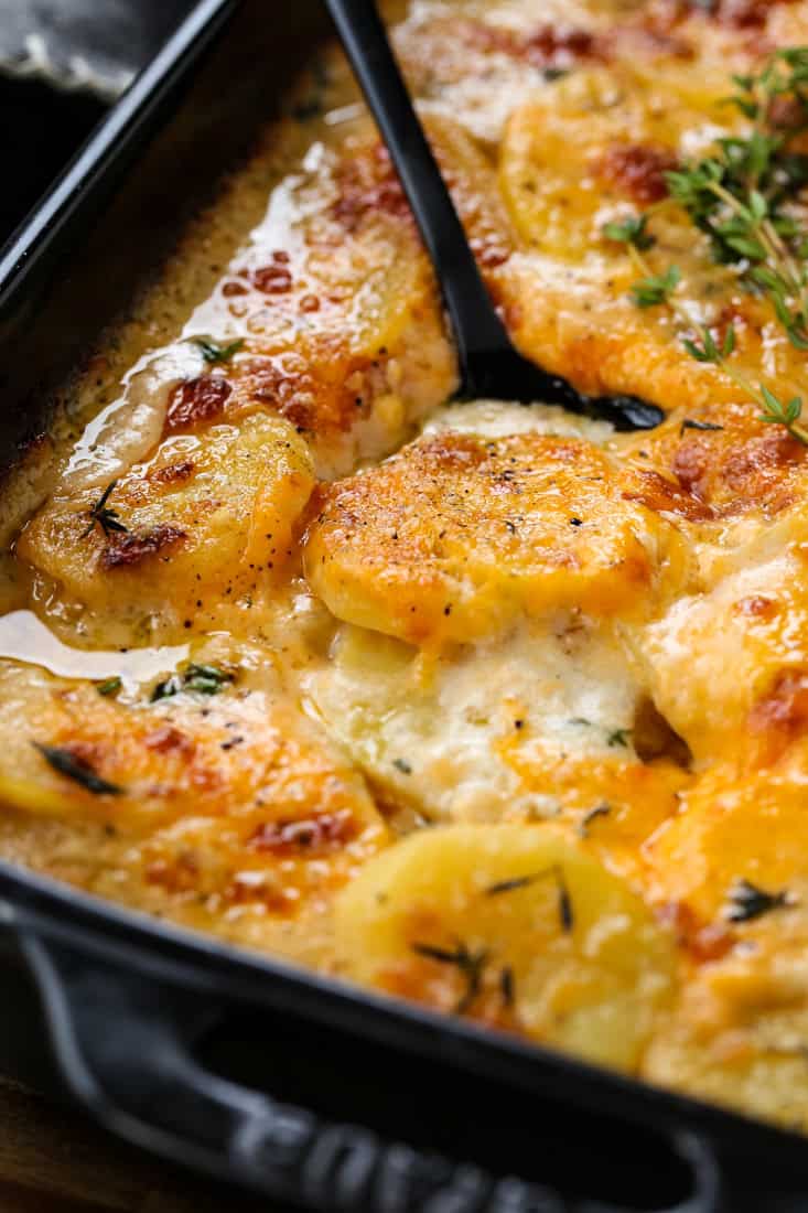 Cheesy Scalloped Potatoes are a favorite holiday side dish! A classic casserole with thinly sliced potatoes in a thick, creamy, cheesy sauce. potato gratin in a black baking dish with a spoon