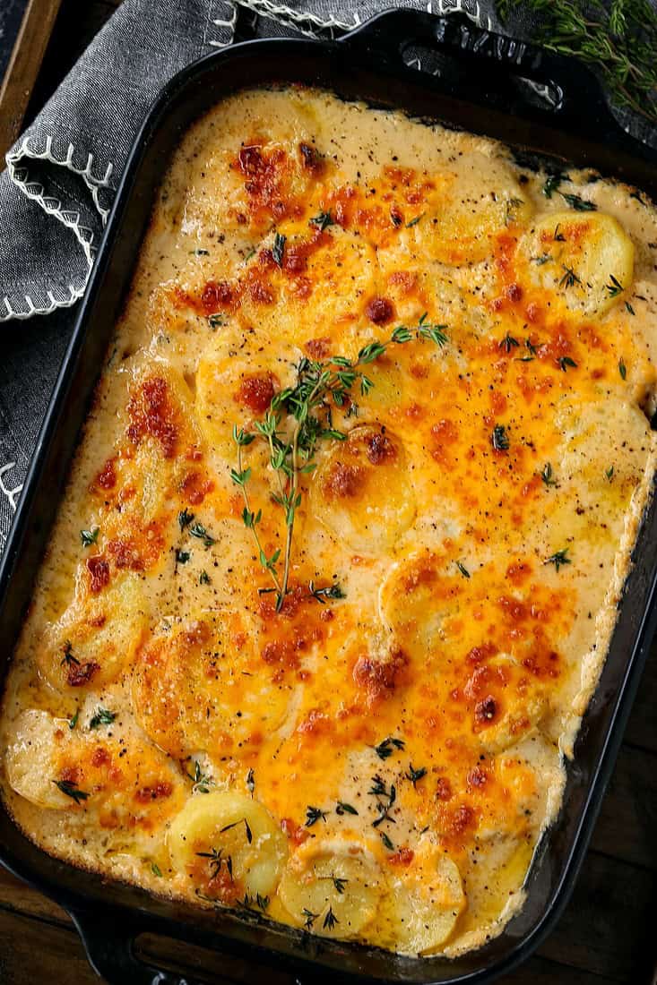 Cheesy Scalloped Potatoes are a favorite holiday side dish! A classic casserole with thinly sliced potatoes in a thick, creamy, cheesy sauce. scalloped potato recipe in a black baking dish with fresh thyme