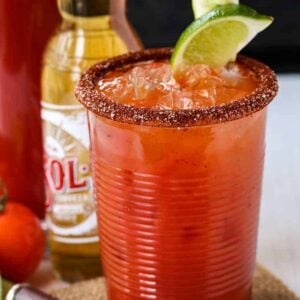 Close up of a Michelada cocktail with a salted rim, garnished with a lime wedge, with a beer bottle and pitcher in the background.