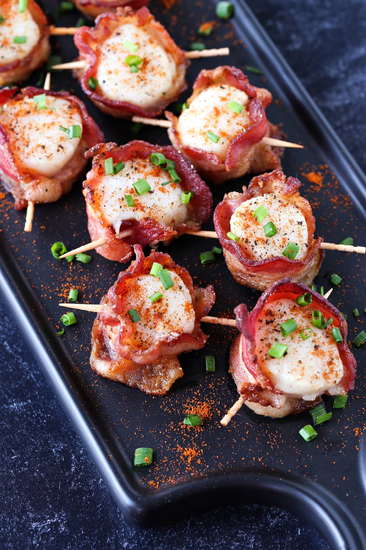 Bacon Wrapped Scallops are a holiday classic appetizer! We've got a hack to make sure that the bacon is actually crispy and not rubbery! bacon wrapped scallops on black serving platter