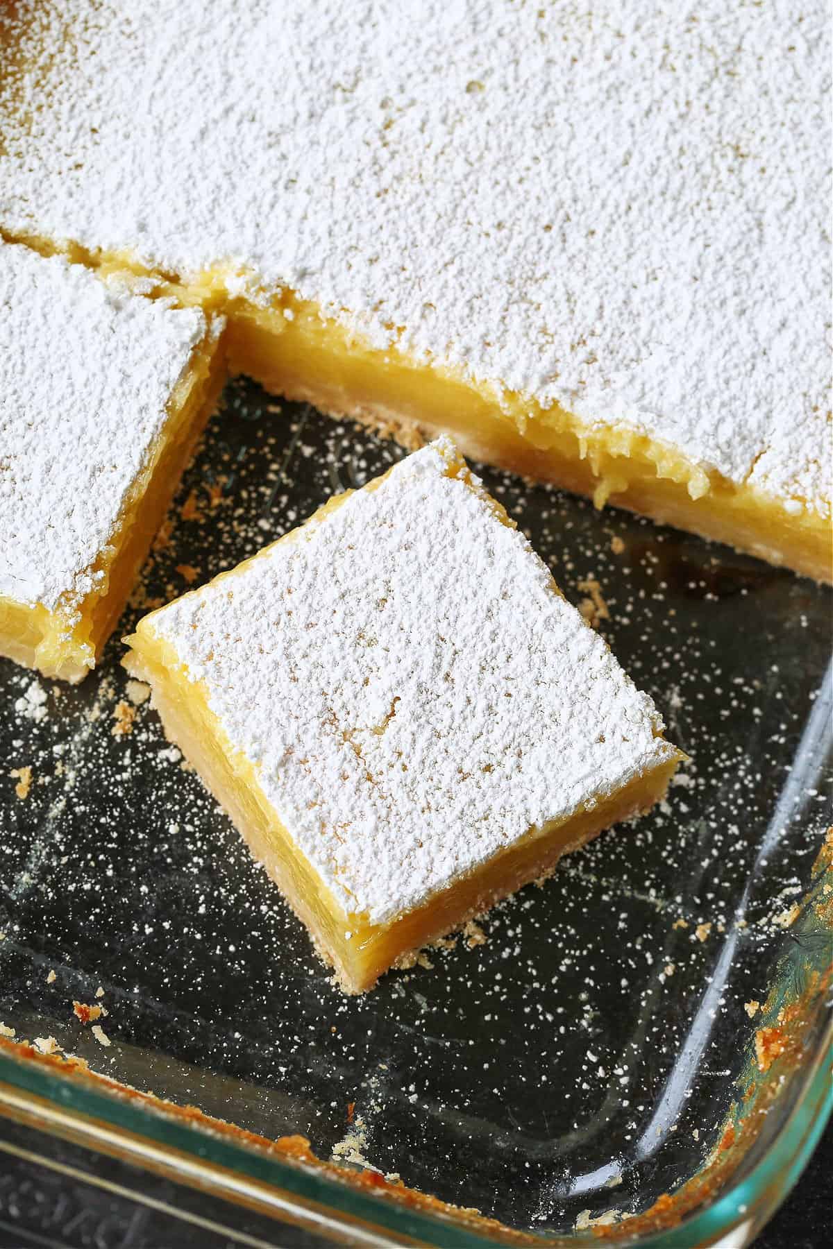 Our Limoncello Lemon Bars are a fun party dessert! Zesty lemon bars with a thick, buttery shortbread topped with a sweet and creamy custard. cut lemon bar in glass dish