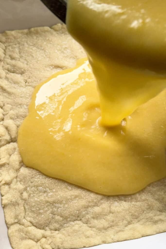 Our Limoncello Lemon Bars are a fun party dessert! Zesty lemon bars with a thick, buttery shortbread topped with a sweet and creamy custard. custard pouring over a baked crust