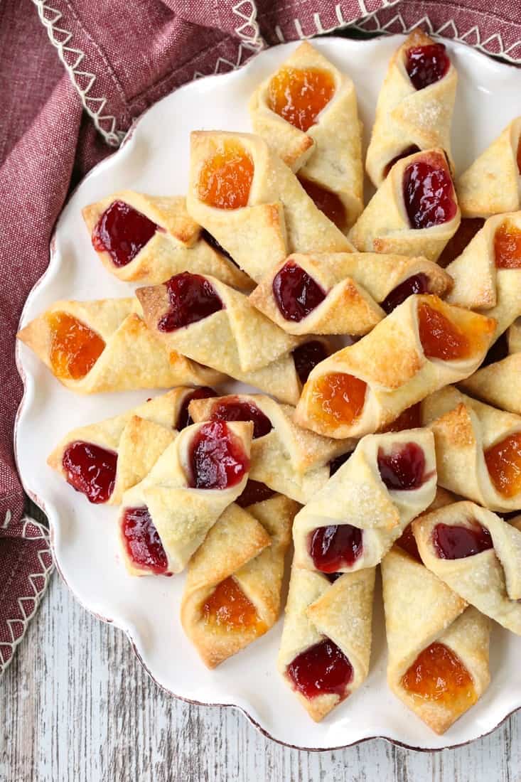 Filled with fruit jelly, Polish Cream Cheese Cookies feature buttery, slightly tangy dough wrapped around apricot and raspberry preserves. Polish Cream Cheese Cookies filled with jelly on a white plate.