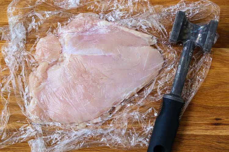 chicken breast on a board with plastic wrap and meat mallet