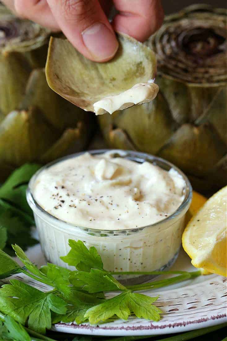 Steamed Artichokes with Garlic Aioli are so easy to make and so good for you! A gorgeous vegetable side dish or appetizer for everyday meals or holiday dinners! Artichoke leaf dipping into garlic aioli with lemon wedge on the side