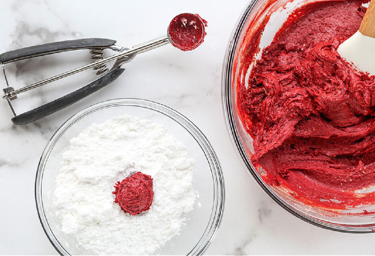 These Red Velvet Crinkle Cookies start off with your favorite boxed cake mix! Soft, cake-like cookies that are perfect for all occasions! cookie scooper and ball of dough in powdered sugar