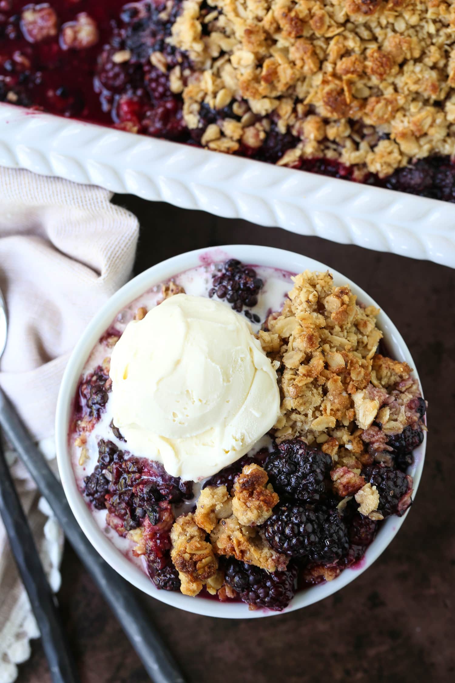 blackberry crispy in a bowl with vanilla ice cream and baking dish in background