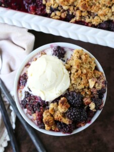 blackberry crisp in a bowl with vanilla ice cream and baking dish in background