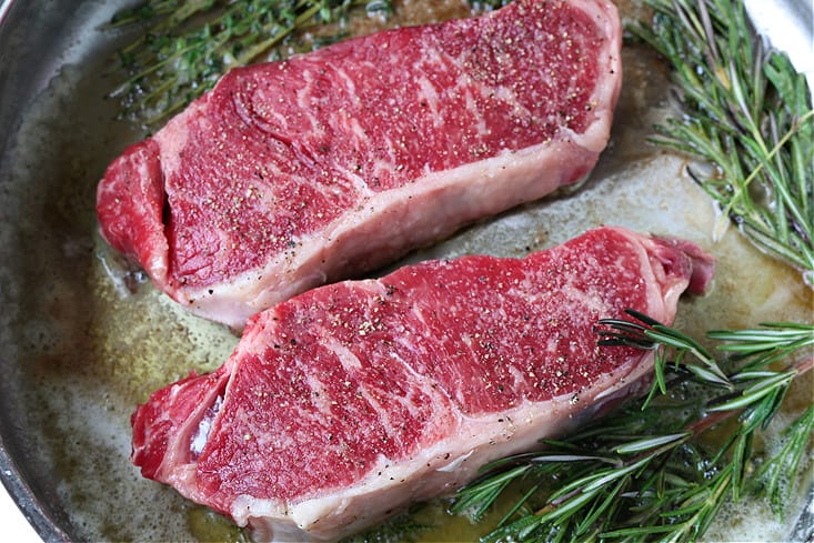 new york strip steaks in a skillet with fresh herbs and butter