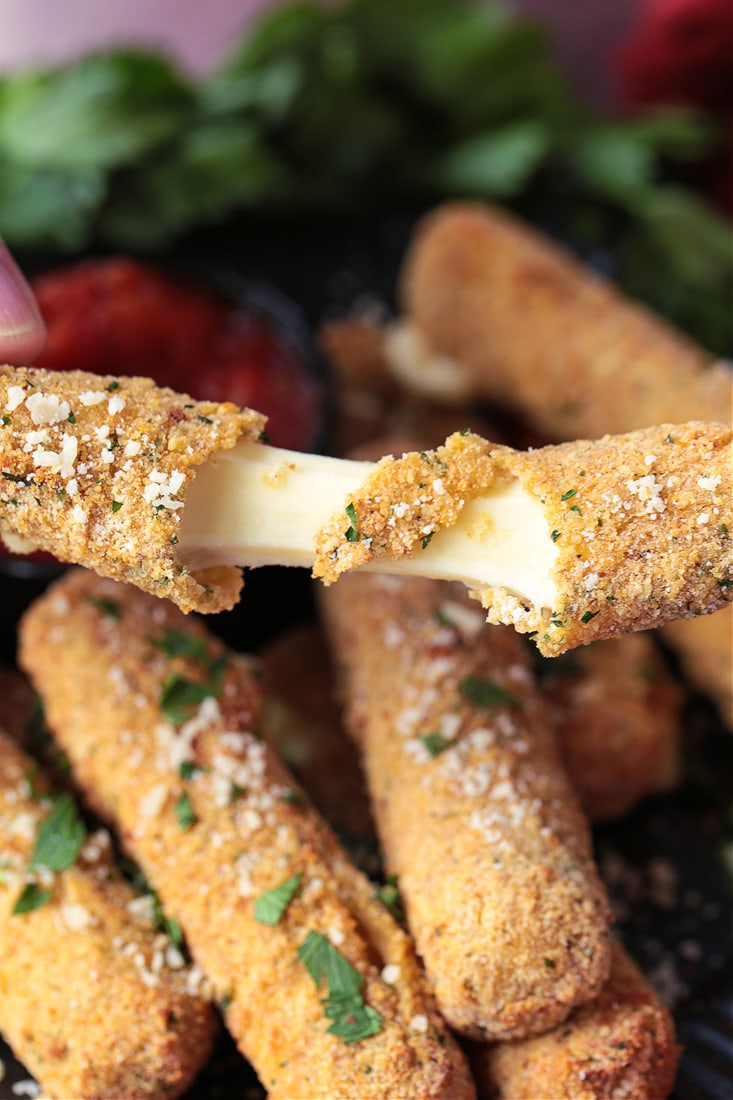 A mozzarella stick being pulled apart with the cheese stretching, with more mozzarella sticks in the background