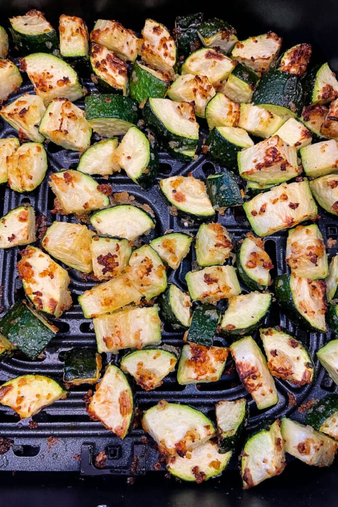 An air fryer basket filled with cooked pieces of zucchini, coated in parmesan cheese and panko breadcrumbs.